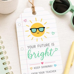 May include: A white gift tag with a sun wearing sunglasses and the text "YOUR FUTURE IS bright KEEP SHINING! FROM YOUR TEACHER". The tag is on a white surface with a pen, sunglasses, and a small plant.