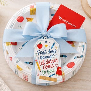 May include: A stack of white paper plates featuring a colorful school supply pattern, tied with a light blue ribbon. A red DoorDash gift card is tucked under the ribbon, and a white tag reads: First days through let dinner come to you!