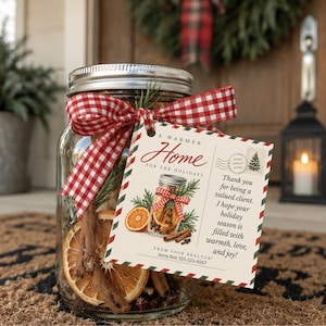 May include: A clear glass mason jar with a silver lid, filled with dried orange slices, cinnamon sticks, and pine sprigs. A red and white gingham ribbon ties a rectangular gift tag featuring the text A WARMER Home FOR THE HOLIDAYS.