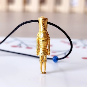 Puede incluir: Un colgante de cascanueces dorado en un collar de cordón negro. El colgante es detallado y tiene una cuenta azul en el cordón.