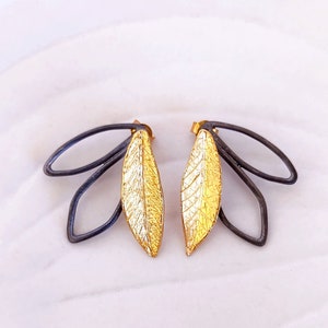 Puede incluir: Un par de pendientes en forma de hoja de oro y negro. Las hojas son texturizadas y tienen un diseño delicado y orgánico. Los pendientes están hechos de metal y tienen un estilo minimalista.