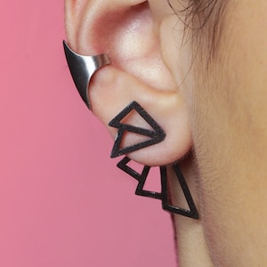 May include: A black ear cuff with a geometric design. The cuff is made of metal and has a sharp, angular shape. It is worn on the upper part of the ear, just above the earlobe.