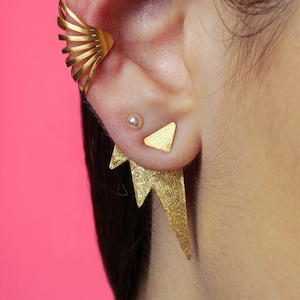 May include: Gold-colored ear cuff and earrings set. The ear cuff features a fan-like design, while the earrings have a triangle stud and a jagged, lightning bolt-shaped drop. The jewelry is set against a pink background.
