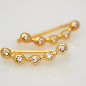 May include: A pair of gold ear cuffs with small, clear gemstones set in each curve. The cuffs are curved and have a delicate, minimalist design.