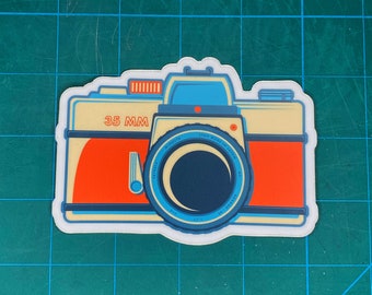 Puede incluir: Una pegatina de cámara de estilo vintage con una lente azul y blanca, un cuerpo rojo y naranja y el texto "35 MM".