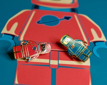 Puede incluir: Dos pines de esmalte con minifiguras Lego en trajes de astronauta. El astronauta de la izquierda lleva un traje rojo con un logotipo blanco en el pecho. El astronauta de la derecha lleva un traje azul con un logotipo blanco en el pecho. Ambos pines están sobre un fondo rojo.
