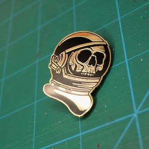 Könnte beinhalten: Goldfarbene Emaille-Anstecknadel mit einem schwarz-weißen Totenkopf in einem Raumanzug. Die Anstecknadel hat ein detailliertes Design und eine glänzende Oberfläche.