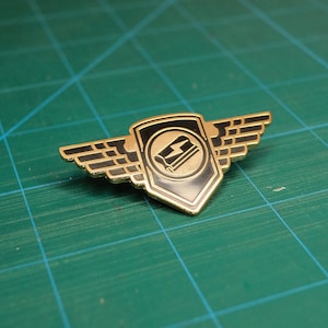 Könnte beinhalten: Goldfarbener Metallstift mit einem schwarzen schildförmigen Design. Der Schild hat eine schwarz-weiße Grafik eines Werkzeugs mit einem Blitzsymbol darin. Der Schild wird von zwei goldenen Flügeln flankiert.