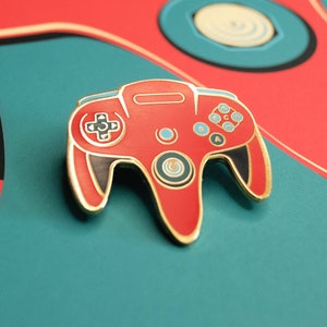 Può includere: Uno spillo a forma di controller Nintendo 64 rosso e dorato. Il controller ha un design in bianco e nero con sfondo rosso. Lo spillo è un oggetto da collezione popolare per gli appassionati di videogiochi.