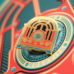 Pode incluir: Um alfinete de esmalte dourado em forma de rádio vintage. O alfinete é vermelho com detalhes em preto e azul, apresentando um design detalhado da face e do mostrador do rádio. O alfinete está sobre um fundo correspondente com o nome "HOROVITZ".