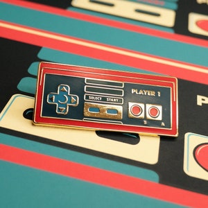 May include: A gold-colored enamel pin shaped like a vintage video game controller. The pin has red and blue accents, and the text "PLAYER 1" is printed on the top. The pin has buttons labeled "SELECT", "START", "B", and "A".