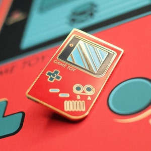 Könnte beinhalten: Eine rote und goldene Emaille-Anstecknadel in Form einer Retro-Handheld-Spielkonsole. Die Anstecknadel hat einen blauen und schwarzen Bildschirm mit dem Text "GAME TOY" auf der Vorderseite.