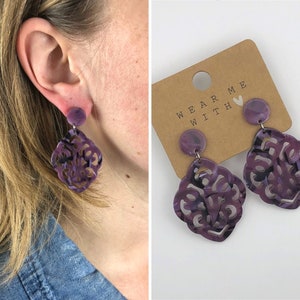 Op de afbeelding: Paarse statement oorbellen met een filigraan ontwerp. De oorbellen bevinden zich op een bruine kaart met de tekst "Wear Me With" en een hart.