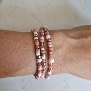 Op de afbeelding: Een armband met drie strengen gemaakt van kleine, ronde kralen in afwisselende kleuren van mat crème, metallic roségoud en dekkend oudroze. De kralen zijn geregen op een flexibel koord, wat zorgt voor een gelaagde uitstraling om de pols.