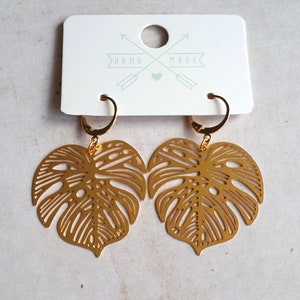 Op de afbeelding: Gouden oorbellen met een uitgesneden monstera-bladontwerp. De oorbellen zijn handgemaakt en hebben een haaksluiting.