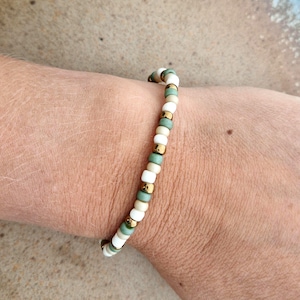 Könnte beinhalten: Ein Perlenarmband mit einem sich wiederholenden Muster aus kleinen, runden Perlen in mattem Weiß, Salbeigrün und Metallic-Gold. Die Perlen sind auf einer flexiblen Schnur aufgereiht und bilden ein einfaches, gleichmäßiges kreisförmiges Design.