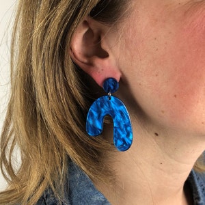 Op de afbeelding: Blauwe en witte marmeren hars oorbellen met een gebogen, abstracte vorm. De oorbellen hangen en hebben een kleinere ronde schijf aan de bovenkant.
