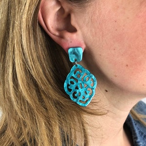 May include: A pair of turquoise dangle earrings featuring a square stud top and a larger, ornate, openwork pendant with a swirling, marbled texture. The earrings are crafted from a smooth, lightweight material with a glossy finish and intricate cutouts.