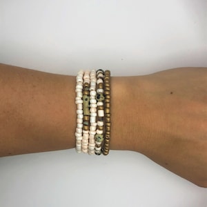May include: A set of seven bracelets stacked on a wrist. The bracelets are made of small beads in white, gold, and brown colours.
