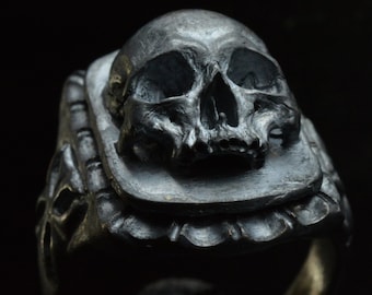 Könnte beinhalten: Ein silberner Totenkopfring mit filigranen Details. Der Totenkopf ist auf einem quadratischen Sockel mit einem dekorativen Rand angebracht. Der Ringband ist dick und hat eine glatte Oberfläche.