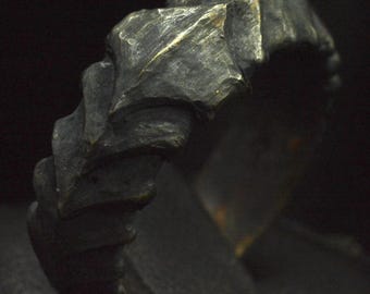 May include: A dark, oxidised metal ring featuring a rugged, textured surface with repeating angular, scale-like protrusions. The band has a weathered, matte finish with subtle highlights of metallic gold showing through the deep charcoal grey patina.