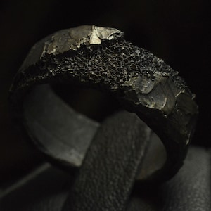 Op de afbeelding: Een zwarte ring gemaakt van ruw, getextureerd metaal. De ring heeft een unieke, organische vorm en een matte afwerking.