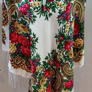 May include: A white tunic with a V-neckline featuring a vibrant floral pattern in red, yellow, and green with paisley accents. The garment has long sleeves and a hemline finished with white fringe, displayed on a mannequin torso.