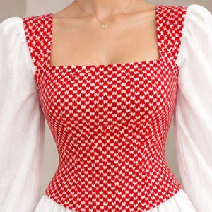 May include: A long-sleeved top featuring a red and white houndstooth patterned bodice with a square neckline and solid white puff sleeves. The fitted bodice transitions into a flared white skirt, creating a structured silhouette with contrasting textures and colors.