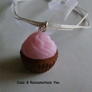 Puede incluir: Un charm de cupcake rosa y marrón en un collar de cinta blanca. El cupcake tiene glaseado rosa y una base marrón.