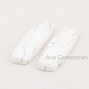 May include: Two white cabochon stones with gray veining. The stones are rectangular and have a smooth, polished surface. Ace Gemstones is printed below the stones.