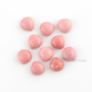Pode incluir: Uma coleção de dez cabochões de opala rosa, cada um com um padrão e forma únicos. Os cabochões estão dispostos em um grupo solto sobre um fundo branco.