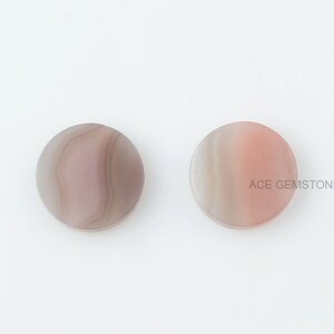 May include: Two round, flat, agate cabochons. The cabochon on the left is a light gray with brown and pink stripes. The cabochon on the right is a light pink with white and gray stripes.  ACE GEMSTONES is printed on the right cabochon.