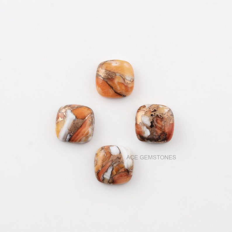 K&ouml;nnte beinhalten: Vier quadratisch geschliffene Edelsteine mit einer Mischung aus orange, wei&szlig;en und braunen Farbt&ouml;nen und goldfarbenen Adern. Die Edelsteine sind auf einer wei&szlig;en Oberfl&auml;che angeordnet. Der Text "ACE GEMSTONES" ist sichtbar.