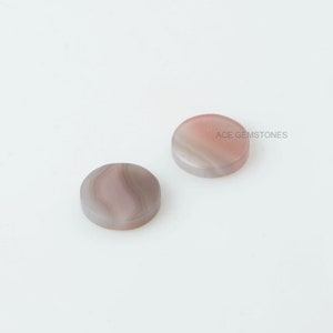 May include: Two round, flat, white and pink agate cabochons with a swirling pattern. ACE GEMSTONES is printed on the white cabochon.