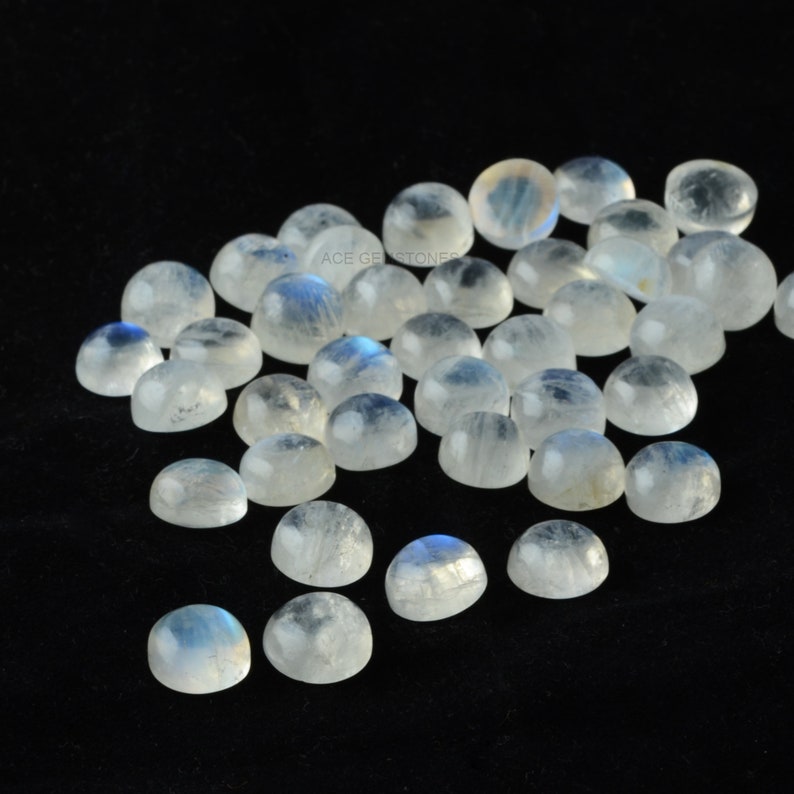 Op de afbeelding: Een verzameling ronde, witte maansteen cabochons met een blauwe, iriserende glans. De stenen zijn verspreid over een zwart oppervlak.