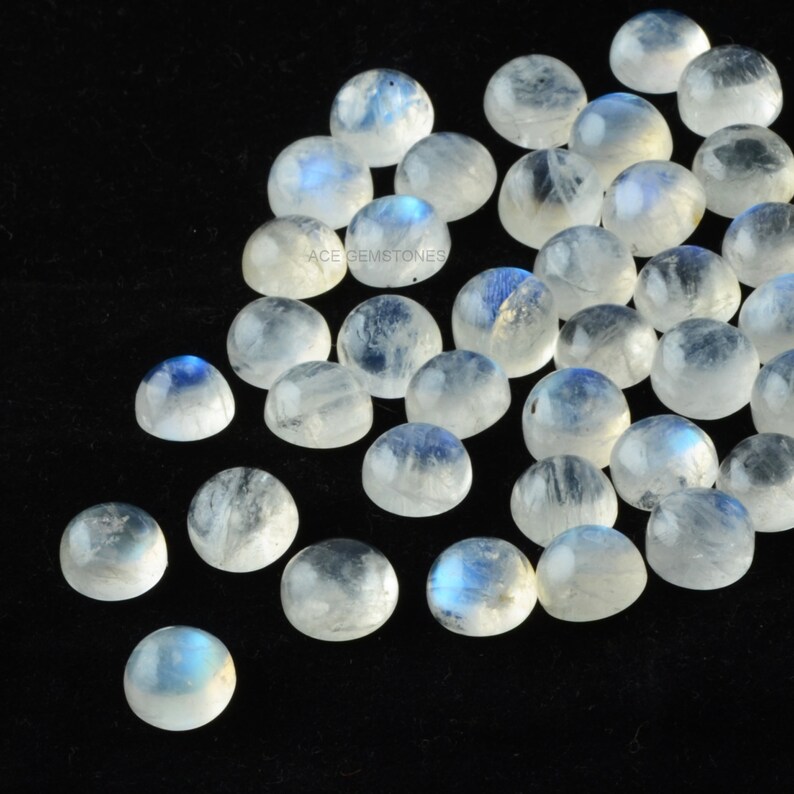 Op de afbeelding: Een verzameling ronde, witte maansteen cabochons met een blauwe, iriserende glans. De stenen zijn verspreid over een zwarte achtergrond.