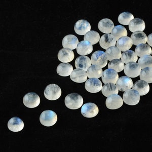 Op de afbeelding: Een verzameling ronde, witte maansteen cabochons met een blauwe, iriserende glans. De stenen zijn verspreid over een zwart oppervlak.  ACE Gemstones is afgedrukt op de afbeelding.