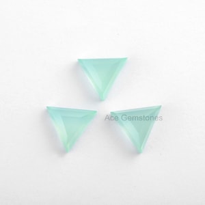 Könnte beinhalten: Drei dreieckige, hellblaue Edelsteinkabochons auf weißem Hintergrund. Ace Gemstones ist in schwarzer Schrift unter den Cabochons gedruckt.