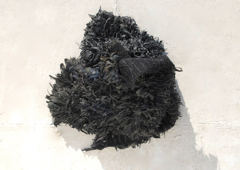 May include: A black, shaggy, textile sculpture made from recycled materials. The sculpture is made of long, thin strips of fabric that are woven together to create a three-dimensional form.