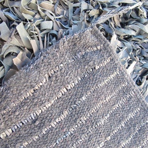 May include: A close-up of a gray woven rug with a textured surface. The rug is made of strips of leather that are woven together to create a striped pattern.