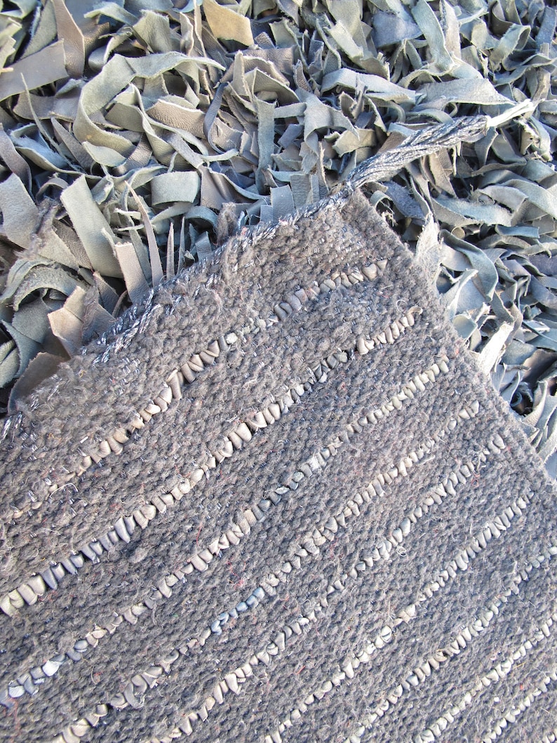 May include: A close-up of a gray woven rug with a textured surface. The rug is made of strips of leather that are woven together to create a striped pattern.
