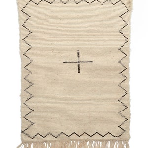 May include: A cream-colored woven rug with a black geometric design. The rug has a fringe border and a simple cross pattern in the center.
