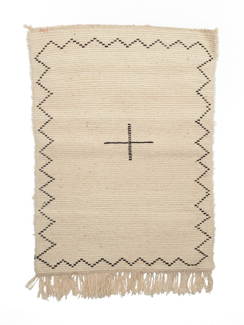 May include: A cream-colored woven rug with a black geometric design. The rug has a fringe border and a simple cross pattern in the center.