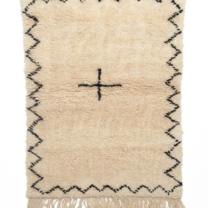 May include: A cream-colored rug with a black border and a black plus sign in the center. The rug has a fringe on the bottom.