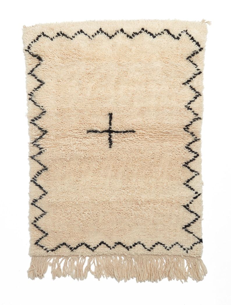 May include: A cream-colored rug with a black border and a black plus sign in the center. The rug has a fringe on the bottom.