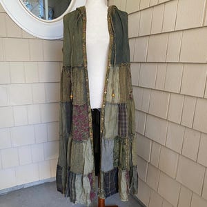 May include: A sleeveless, knee-length hooded vest featuring a patchwork design of various green, brown, and dark purple fabrics. The garment includes a button-down front closure, a hood with drawstring ties, and a multi-textured, layered construction.