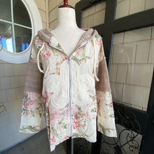 May include: A floral print zip-up hoodie with a cream-colored base, featuring pink and green floral patterns. The hoodie has a cream-colored flower applique on the front, with striped sleeves and a drawstring hood. The jacket is displayed on a mannequin.