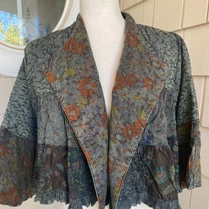 May include: A cropped jacket featuring a patchwork design with various floral patterns in shades of green, brown, and orange. The garment has long sleeves, a wide lapel collar, and a ruffled hemline, constructed from multiple textured fabric panels.