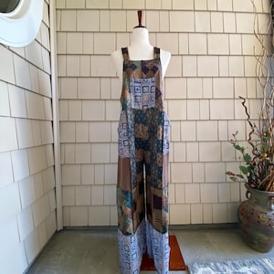 May include: A full-length jumpsuit featuring a patchwork design with various geometric and floral patterns in shades of brown, blue, and white. The garment has wide legs, a square neckline, and thick shoulder straps, displayed on a mannequin.