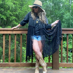 May include: A long black denim jacket featuring a lace overlay, a straw hat with a black band, a dark t-shirt with a gold heart graphic, light blue denim shorts, and tan leather cowboy boots with white floral embroidery and stitching details.